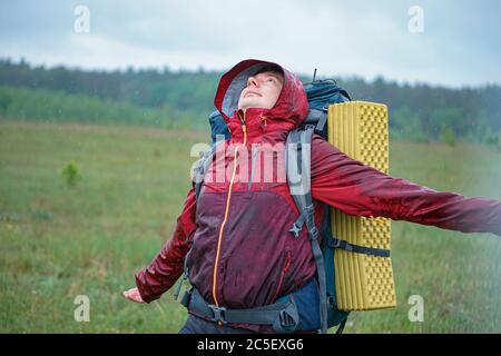 Touristenreisender in einer Membranjacke steht unter einem Regensturm in den Bergen. Regentropfen fallen Regenmantel auf Gesicht und Kleidung Stockfoto