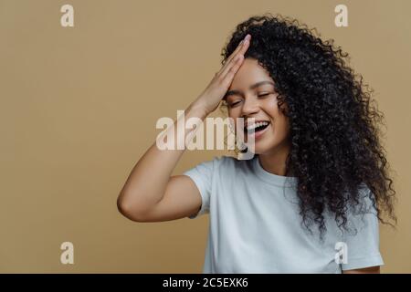 Horizontale Aufnahme von glücklicher positiver afroamerikanischer Frau hat Spaß, hält die Hand auf der Stirn, kichert glücklich, schließt die Augen, hat buschiges lockiges Haar, trägt Casua Stockfoto