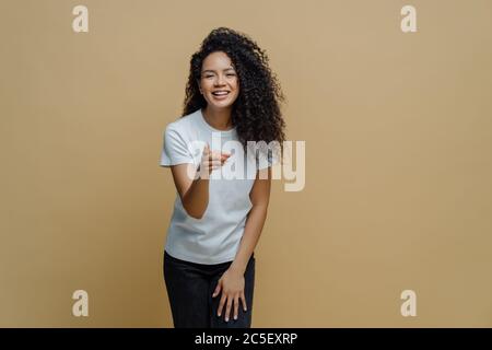 Fröhlich energische junge Frau mit lockigen Afro-Haaren zeigt Zeigefinger nach vorne auf die Kamera, fühlt sich positiv und wählt jemanden, trägt weißes T-Shirt und Stockfoto