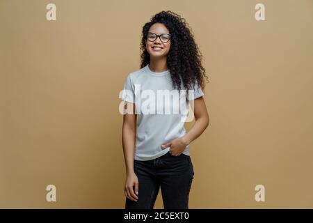 Positive ethnische Schülerin fühlt sich nach der Prüfung frei, lächelt breit, hält die Hand in der Tasche von Jeans, trägt transparente Brille, posiert gegen Stockfoto