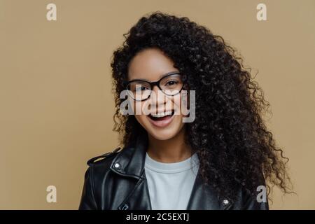 Kopfschuss der fröhlichen afroamerikanischen Frau mit dunklen lockigen Haaren, hat den Ausdruck des erfreulichen Gesichts, das fröhliche Gespräch, trägt die transparente Brille und blac Stockfoto