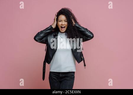 Emotional verrückte Erwachsene Frau greift den Kopf und schreit laut, hört sehr laute Musik, hält den Mund geöffnet, in stilvoller Lederjacke gekleidet, isoliert auf pi Stockfoto