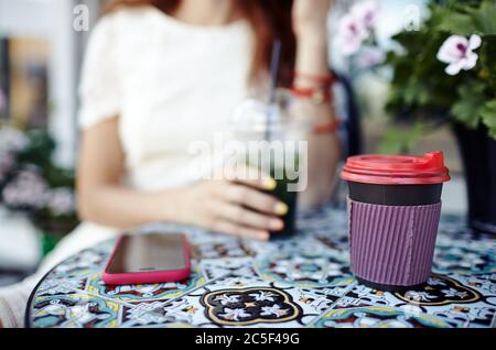 Weibliche Hand hält Plastikbecher mit einem Mojito in einem Stadtcafe. Papiertasse Kaffee und Smartphone auf dem Tisch Stockfoto