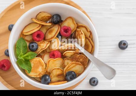 Frühstück weiße Schüssel mit winzigen Pfannkuchen Müsli mit Heidelbeeren, Himbeeren und Minze auf Holzhintergrund, Nahaufnahme, selektive Fokus Stockfoto