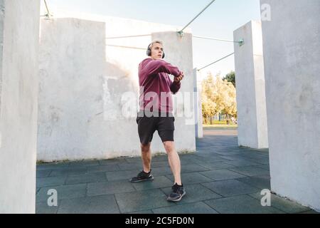 Fitness-Mann wärmt sich im Freien auf dem Sportplatz und hören Sie Musik in Kopfhörern. Dreht den Körper und schwingt die Hände Stockfoto