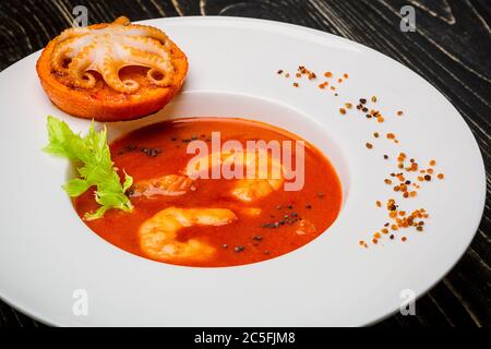 Schüssel Tomatensuppe mit Garnelen mit einem kleinen Oktopus auf einer orangefarbenen Scheibe auf schwarzem Holzgrund gebacken, Draufsicht Stockfoto