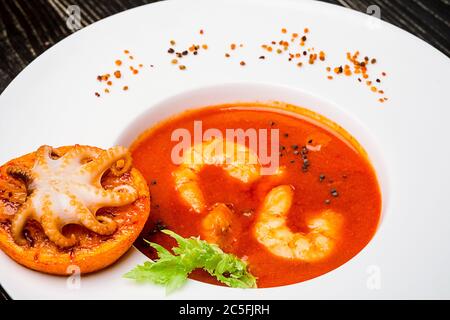 Schüssel Tomatensuppe mit Garnelen mit einem kleinen Oktopus auf einer orangefarbenen Scheibe auf schwarzem Holzgrund gebacken, Draufsicht Stockfoto