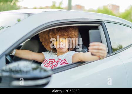 Frau mit afro Haar, die ein Foto mit ihrem Smartphone neben ihrem Auto Stockfoto
