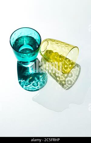 Ein Kristallglas fällt und hinterlässt eine Pfütze Wasser unter dem starken Sonnenlicht, das bunte, strukturierte Schatten auf einen weißen Tisch wirft. Kreative Fotografie Stockfoto