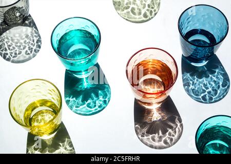 Set von farbigen Glas Tassen mit Wasser gefüllt, die unter einem starken Licht, das seine strukturierten und farbigen Schatten auf einen weißen Tisch projiziert leuchten. Creativ Stockfoto