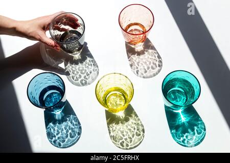 Person nimmt eine der farbigen Glas Tassen unter dem starken Sonnenlicht durch das Fenster und wirft seine strukturierten, farbigen Schatten auf einem weißen Stockfoto