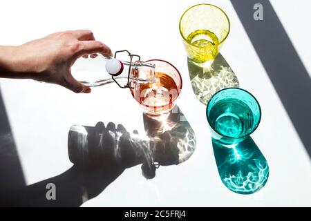 Eine Person gießt Wasser in farbige Glasbecher unter dem starken Sonnenlicht, das durch das Fenster eintritt und wirft seine strukturierten, farbigen Schatten auf ein Whi Stockfoto