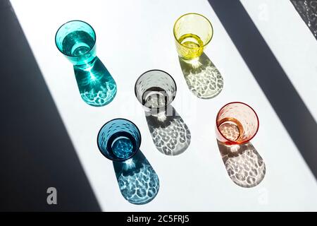 Ein Satz von farbigen Glasbechern, gefüllt mit Wasser unter einem Fenster, durch das Sonnenlicht eindringt und seine strukturierten, farbigen Schatten auf einen weißen Tisch wirft. Stockfoto