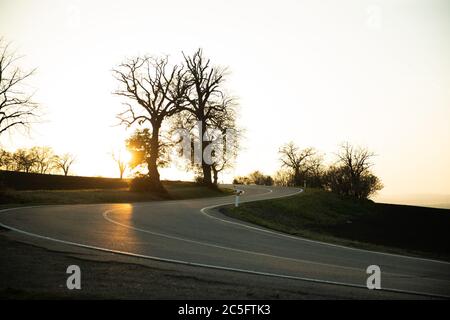 Eine kurvenreiche Straße in der Region Südmähren bei Sonnenuntergang mit Strahlen hüpfend von der Asphalt vor dem Hintergrund von ein paar Bäumen und einem orangen Himmel mit Stockfoto