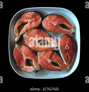 Lachssteaks liegen auf einer weißen quadratischen Keramikplatte. Roter Fisch auf einem isolierten schwarzen Hintergrund Stockfoto