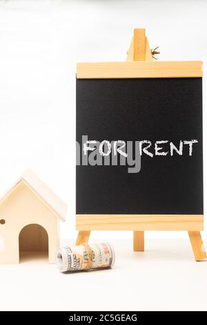„FOR RENT“-Formulierungen auf einer Tafel mit zusammengerolltem Geld und Miniaturhaus Stockfoto