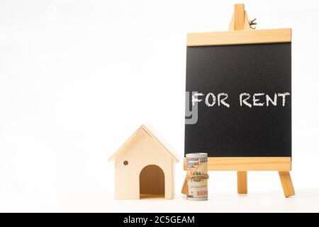 „FOR RENT“-Formulierungen auf einer Tafel mit zusammengerolltem Geld und Miniaturhaus Stockfoto