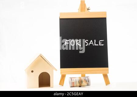 „ZUM VERKAUF“-Formulierungen auf einer Tafel mit zusammengerolltem Geld und Miniaturhaus Stockfoto