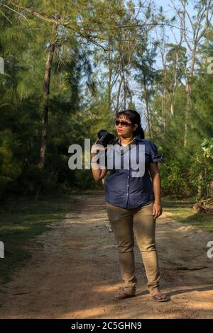 Ein weibliches Modell, das auf einer Waldstraße mit Kamera in der Hand steht, verwischt den selektiven Fokus im Hintergrund Stockfoto