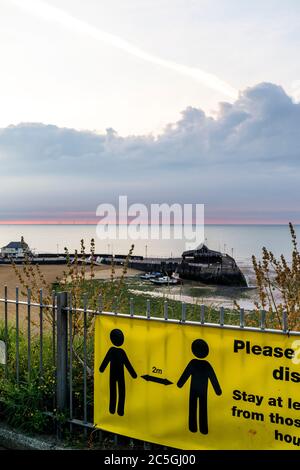 Dawn bricht über Broadstairs Hafen und die Nordsee. Dünne Linie des roten Himmels am Horizont, die sich in graue dunkle Wolken verwandelt. Im Vordergrund Corona Virus, COVID Schilder auf Strandgeländer, re soziale Distanzierung und halten zwei Meter auseinander. Stockfoto