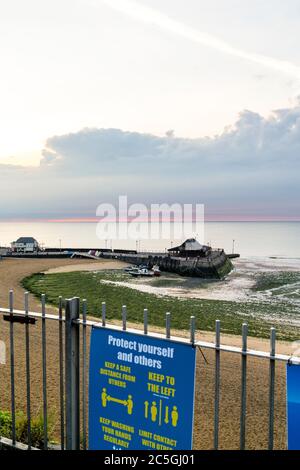 Dawn bricht über Broadstairs Hafen und die Nordsee. Dünne Linie des roten Himmels am Horizont, die sich in graue dunkle Wolken verwandelt. Im Vordergrund Corona Virus, COVID Schilder auf Strandgeländer, re soziale Distanzierung und halten zwei Meter auseinander. Stockfoto