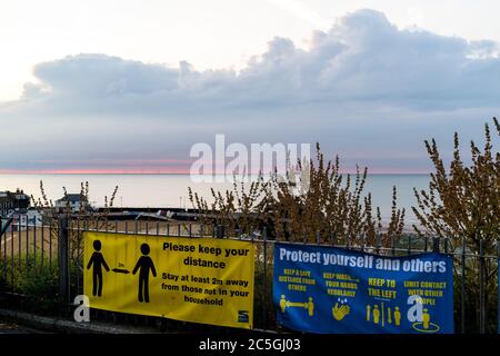 Dawn bricht über Broadstairs Hafen und die Nordsee. Dünne Linie des roten Himmels am Horizont, die sich in graue dunkle Wolken verwandelt. Im Vordergrund Corona Virus, COVID Schilder auf Strandgeländer, re soziale Distanzierung und halten zwei Meter auseinander. Stockfoto