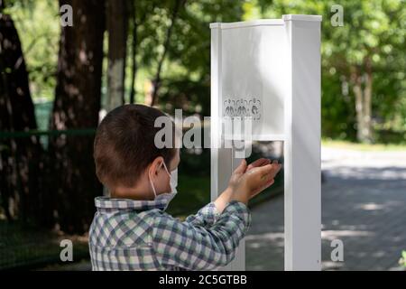 Ein kleiner Junge in einer Schutzmaske wäscht sich gründlich die Hände mit einem Desinfektionsmittel, um sich vor Mikroben an einem öffentlichen Ort im Park zu schützen Stockfoto