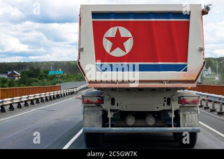 Großer LKW mit der Nationalflagge Nordkoreas, der sich auf der Autobahn bewegt, vor dem Hintergrund der Dorf- und Waldlandschaft. Konzept des Exports Stockfoto
