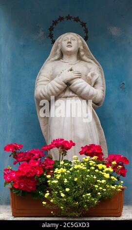 Einfache Statue von Maria mit in einer blau bemalten Nische in einer Wand. Blumenkachtel mit gelben und roten Blüten davor Stockfoto