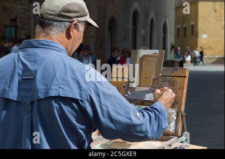 Männlicher Amateur-Künstler in blauem Hemd malt an einem sonnigen Tag ein Haus in einer Straße in Lucca. Das Panel ist auf einer Staffelei Stockfoto