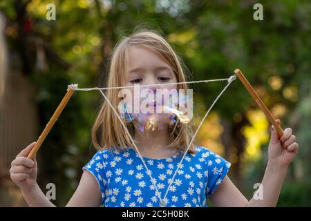 Junges Mädchen in einem blauen und weißen Kleid weht große Blasen mit String in einem Hinterhof Stockfoto