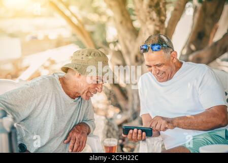Zwei positive lächelnde Silber Surfer Senior Mann verwenden Smartphone und Tee trinken. Aktive gesunde Senioren Spaß im Garten. Stockfoto