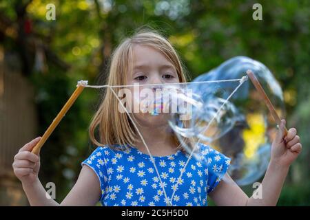 Junges Mädchen in einem blauen und weißen Kleid weht große Blasen mit String in einem Hinterhof Stockfoto