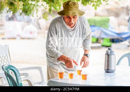 Gesunder älterer Mann, der heißen Tee aus der Teekanne in kleine Teegläser in einem Garten gießt. Stockfoto