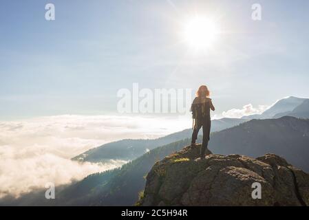 Frau auf Berggipfel ruhen, Blick auf die dramatische Landschaft Wolken über dem Tal, klaren blauen Himmel Sonnenstrahlen im Hintergrund. Sommeraktivitäten fitnes Stockfoto