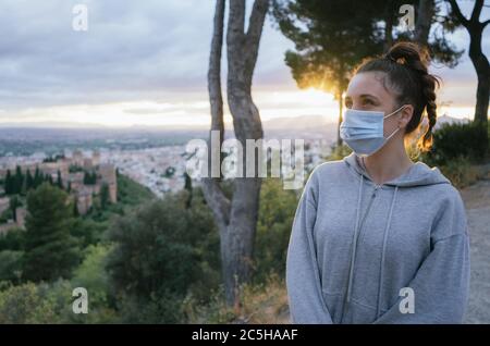 Junge Mädchen mit einer Maske von der covid, Verschmutzung mit der Sonne im Hintergrund, über der Stadt der Alhambra, Spanien. Sommeruntergang. Stockfoto