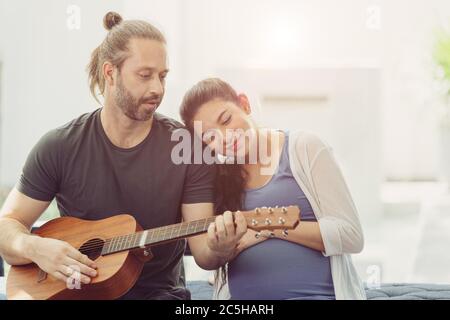 Ehemann mit schwangeren Frau zu Hause bleiben schön spielen Gitarre Musik für Baby küssen und zeigen Liebe zusammen Stockfoto