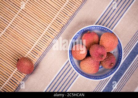 Litschi in einer traditionellen chinesischen blau-weißen Schale mit Bambusmatte und gestreifter Tischdecke Stockfoto