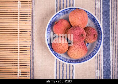 Litschi in einer traditionellen chinesischen blau-weißen Schale mit Bambusmatte und gestreifter Tischdecke Stockfoto