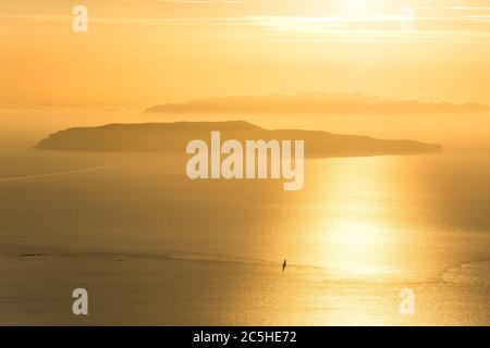 Kleine Insel und einsames Segelschiff auf dem Meer während dramatischen goldenen Sonnenuntergang Stockfoto