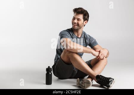 Lächelnder junger Sportler sitzt isoliert auf grauem Hintergrund, ruht sich nach dem Training aus, schaut weg Stockfoto