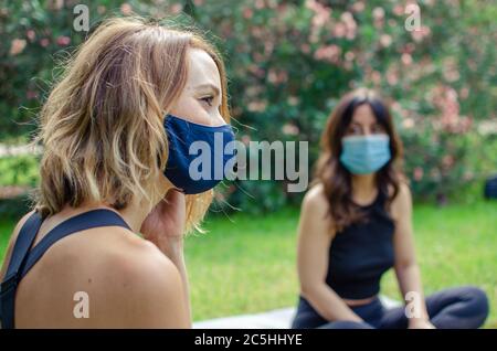Zwei Frauen im Park mit Gesichtsmasken. Sie sitzen auf dem Gras Stockfoto
