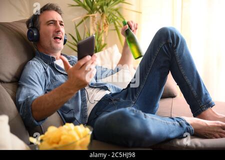 Fröhlicher Mann, der zu Hause auf dem Sofa singt und einen Snack auf dem Sofa nimmt, während er mit Kopfhörern von einem Handy Musik hört Stockfoto