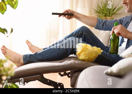 Mann, der Fernsehen schaut, sitzt bequem auf einem Sofa und hat einen Snack mit Bier und eine Fernbedienung in den Händen. Stockfoto