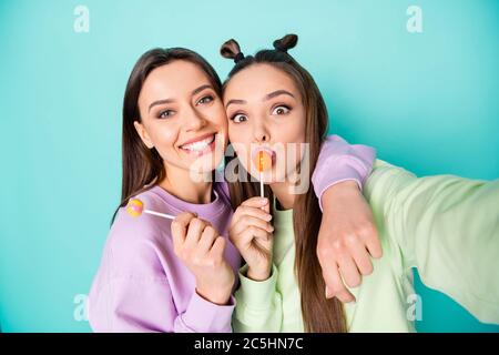 Foto von zwei lustigen Damen halten Lollipop chupa chups Hände kindisch lecker Süßigkeiten machen Selfies tragen grün violett Pullover isoliert pastellgrün teal Farbe Stockfoto