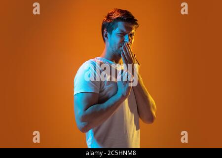 Verungustet. Kaukasischer junger Mann Porträt auf Gradienten orange Studio Hintergrund in Neonlicht. Schönes männliches Modell in weißem Hemd. Konzept der menschlichen Emotionen, Gesichtsausdruck, Verkauf, Anzeige. Copyspace. Stockfoto