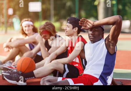 Erschöpfter schwarzer Basketballspieler, der sich mit seinem vielfältigen Team auf dem Sportplatz draußen ausruhte Stockfoto