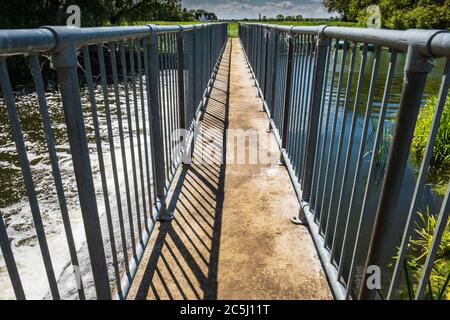 Hoher Kontrast, flacher Fokus einer schmalen Fußgängerbrücke, die im Frühsommer ein großes Wehr in der englischen Landschaft überquert. Stockfoto