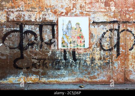 Varanasi, Uttar Pradesh, Indien : Nahaufnahme einer verwitterten Wand mit Devanagari-Schrift und einer Fliese, die die Götter Shiva, Parvati und Ganesh darstellt. Stockfoto