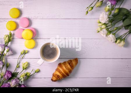Makronen und Croissant mit Blumen und Tasse Kaffee auf weißem Holzhintergrund mit Kopierfläche. Draufsicht Stockfoto